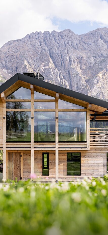 Uno chalet a due piani con una facciata in legno invecchiato in mezzo a un prato, con la catena montuosa del Latemar sullo sfondo. | © Meraner & Hauser