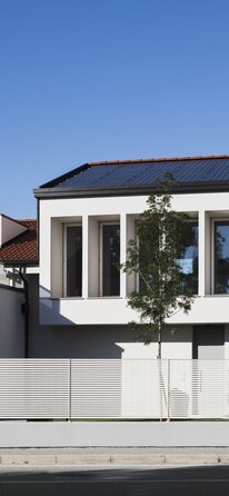 A two-storey residential building with a large row of windows on the upper floor and photovoltaic system on the roof | © Davide Perbellini