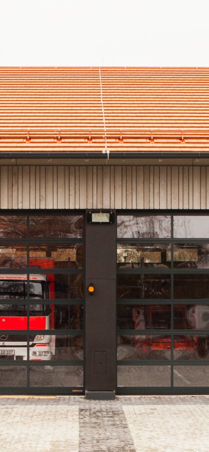 Einfahrt einer Feuerwehrhalle mit Außenschalung aus Holz und Einfahrtstoren aus Glas. | © Regina Sedlmayer