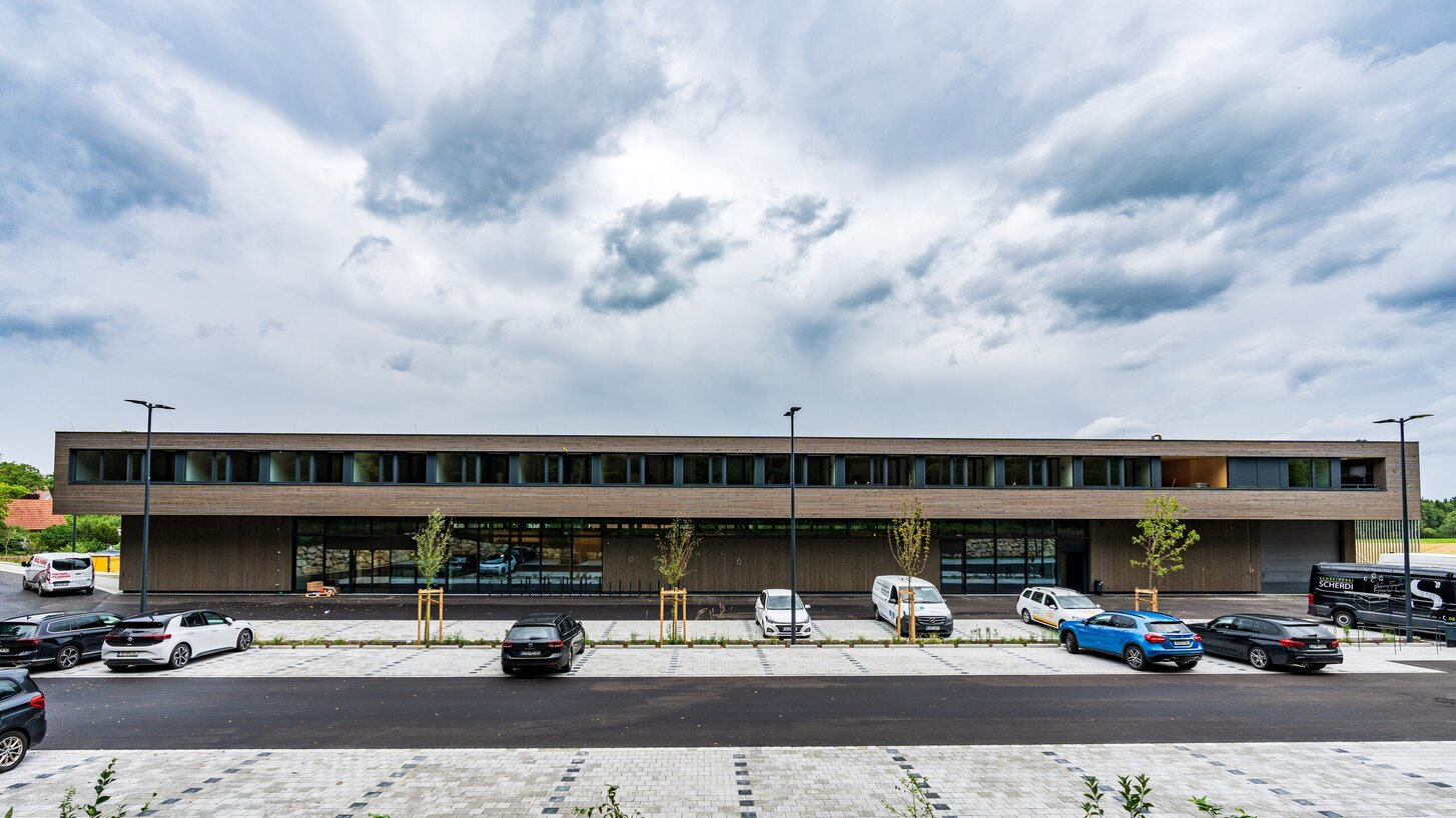A large two-storey building with a façade clad in wooden strips and a car park in front of it | © Roland Wehinger