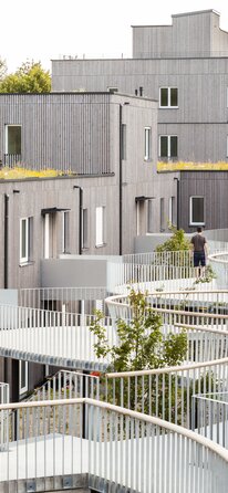 Un grande complesso residenziale con edifici di tre e quattro piani con facciate in listelli di legno impregnati in grigio e un pergolato curvo come ingresso. | © Regina Sedlmayer
