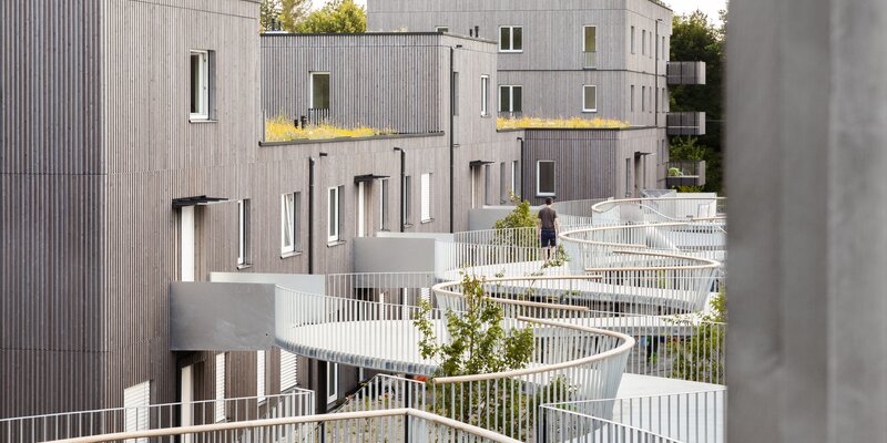 Eine große Wohnanlage mit drei- und vierstöckigen Gebäuden mit Fassaden aus vorvergrauten Holzleisten und einem geschwungenen Laubengang als Zugang | © Regina Sedlmayer
