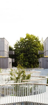 Un grande complesso residenziale con edifici di tre e quattro piani con facciate in listelli di legno impregnati in grigio e un pergolato curvo come ingresso. | © Regina Sedlmayer