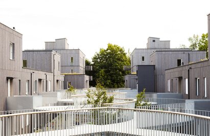 A large residential complex with three and four-storey buildings with façades made of pre-greyed wooden slats and a curved pergola as an entrance | © Regina Sedlmayer