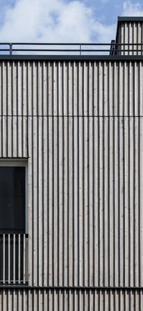 La facciata di un edificio con rivestimento di facciata con listelli in legno impregnati color grigio | © Regina Sedlmayer
