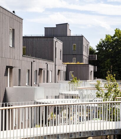 Un grande complesso residenziale con edifici di tre e quattro piani con facciate in listelli di legno impregnati in grigio e un pergolato curvo come ingresso. | © Regina Sedlmayer
