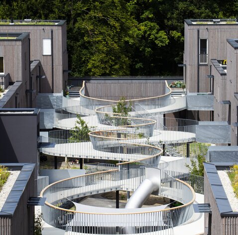 Un grande complesso residenziale con edifici di tre e quattro piani con facciate in listelli di legno impregnati in grigio e un pergolato curvo come ingresso. | © Regina Sedlmayer