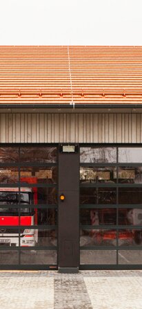 Einfahrt einer Feuerwehrhalle mit Außenschalung aus Holz und Einfahrtstoren aus Glas. | © Regina Sedlmayer