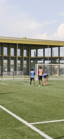 Ein Fußballfeld mit Spielern, im Hintergrund ein großes Gebäude mit vielen Fenstern und einem goldig glänzenden Dach | © Michele Nastasi