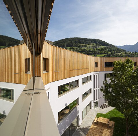 Ein Schulgebäude, auf den drei verputzten Untergeschossen steht ein vierter Stock mit einer vertikal laufenden Verkleidung aus Lärchenholz | © Samuel Holzner