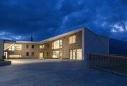 A large building with a white painted exterior wall and wooden cladding on the façade. Light burns inside | © Oliver Jaist