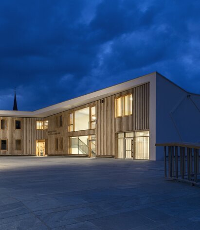 Ein großes Gebäude mit einer weiß gestrichenen Außenwand und Holzverkleidung an der Fassade. Im Inneren brennt Licht | © Oliver Jaist