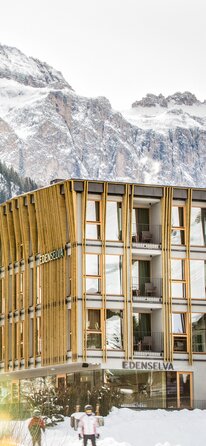 Ein vierstöckiges Hotelgebäude im Winter, die drei Obergeschosse sind mit vertikalen Lamellen verkleidet, im Hintergrund ein Wald und Berge | © Edenselva - Mattia Gasparotto