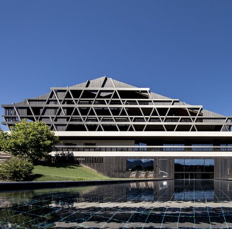 Ein großes Gebäude mit einer Fassade aus dunklem Holz, im Vordergrund ein Freischwimmbad | © Florian Andergassen