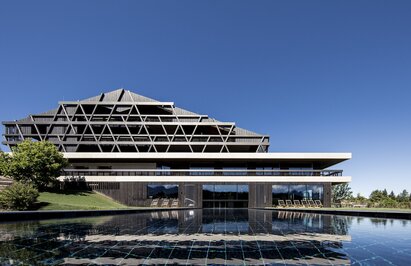 Ein großes Gebäude mit einer Fassade aus dunklem Holz, im Vordergrund ein Freischwimmbad | © Florian Andergassen