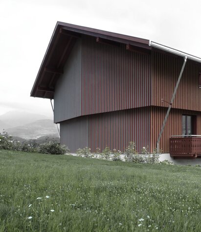 Un grande edificio con una facciata in legno di colore rosso divisa da listelli verticali con sezione triangolare dipinte di giallo da un lato e di blu dall'altro. | © Gustav Willeit