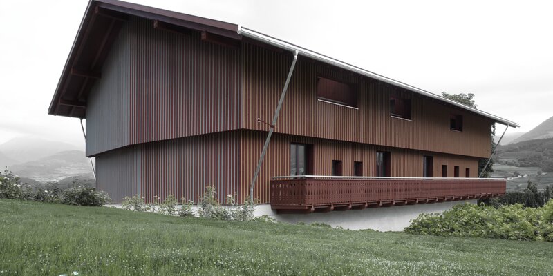 Ein großes Gebäude mit einer rotfarbigen Holzfassade, die durch vertikal verlaufende, dreieckige Leisten gegliedert ist, die auf der einen Seite gelb und auf der anderen Seite blau gestrichen sind | © Gustav Willeit