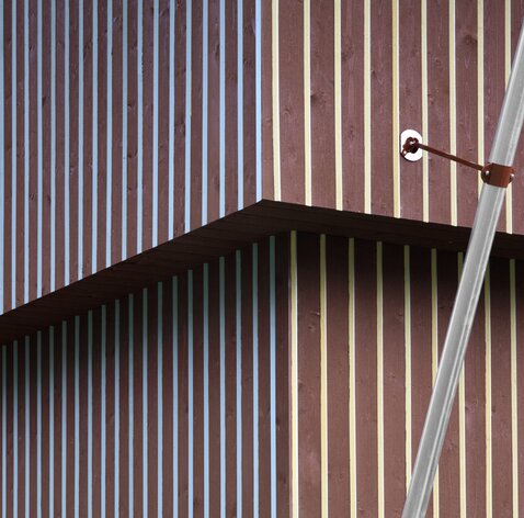 Una facciata in legno di colore rosso divisa da listelli verticali con sezione triangolare dipinte di giallo da un lato e di blu dall'altro. | © Gustav Willeit