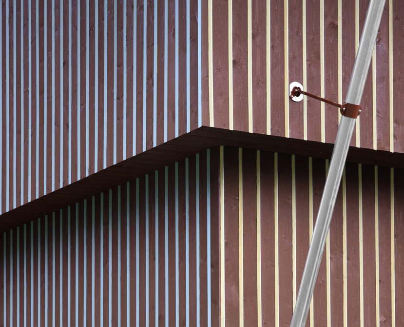 Una facciata in legno di colore rosso divisa da listelli verticali con sezione triangolare dipinte di giallo da un lato e di blu dall'altro. | © Gustav Willeit