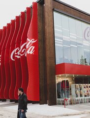 Exterior view of a rectangular wooden pavilion. At the front a large glass façade with the inscription "Coca-Cola", at the side red slats go over the entire height of the building. | © LignoAlp