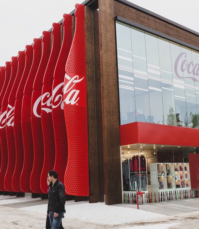 Exterior view of a rectangular wooden pavilion. At the front a large glass façade with the inscription "Coca-Cola", at the side red slats go over the entire height of the building. | © LignoAlp