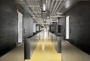 A corridor with a yellow floor leads into a modern-looking building, to the left and right of which are walls made of black impregnated wood | © Davide Perbellini