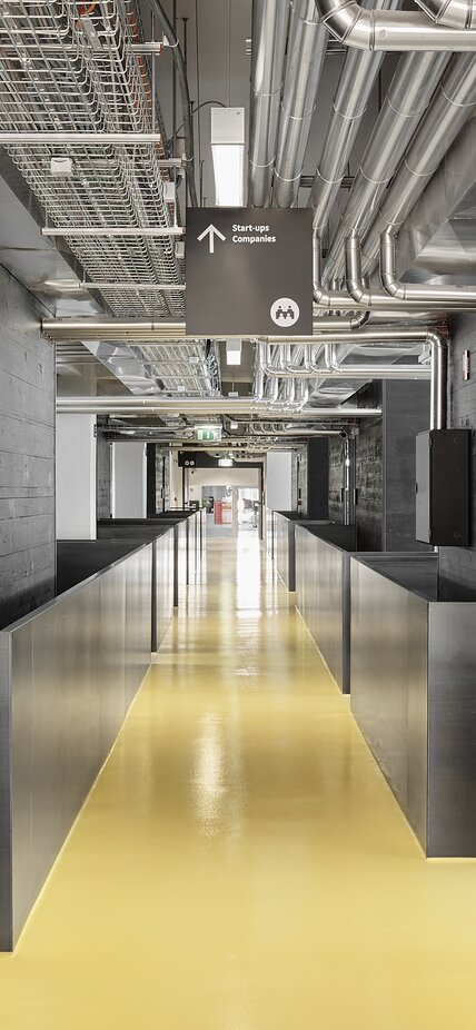 Un corridoio dal pavimento giallo conduce in un edificio dall'aspetto moderno, a sinistra e a destra del quale si trovano dei pareti in legno impregnato nero. | © Davide Perbellini