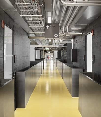 Un corridoio dal pavimento giallo conduce in un edificio dall'aspetto moderno, a sinistra e a destra del quale si trovano dei pareti in legno impregnato nero. | © Davide Perbellini