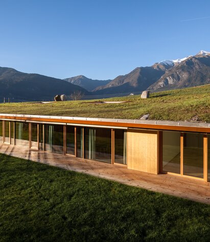 Ein Gebäude, das in den Hang gebaut ist. Sichtbar sind nur eine Fensterfront mit Holzfenstern, das Dach ist begrünt. Berge im Hintergrund. | © Davide Cornacchini