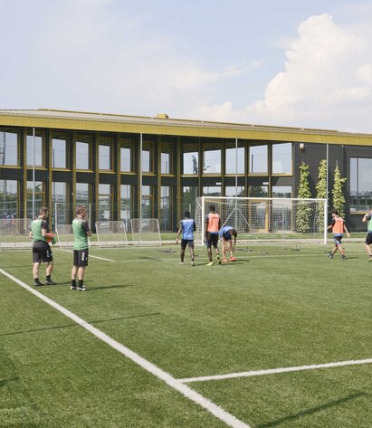 Un campo da calcio con giocatori, sullo sfondo un grande edificio con molte finestre e un tetto color oro lucido | © Michele Nastasi