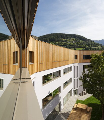 Ein Schulgebäude, auf den drei verputzten Untergeschossen steht ein vierter Stock mit einer vertikal laufenden Verkleidung aus Lärchenholz | © Samuel Holzner