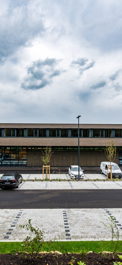 A large two-storey building with a façade clad in wooden strips and a car park in front of it | © Roland Wehinger
