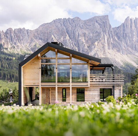 A two-storey chalet with a façade made of old wood in the middle of a meadow, with the Latemar mountain range in the background | © Meraner & Hauser