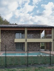 Façade of a two-storey building, partly plastered and coloured yellow and partly clad in stone. The balustrades of the terraces are made of glass, a photovoltaic system can be seen on the roof and a meadow with a swimming pool can be seen in the foreground | © Davide Perbellini