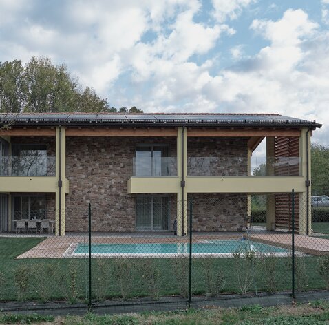 Façade of a two-storey building, partly plastered and coloured yellow and partly clad in stone. The balustrades of the terraces are made of glass, a photovoltaic system can be seen on the roof and a meadow with a swimming pool can be seen in the foreground | © Davide Perbellini